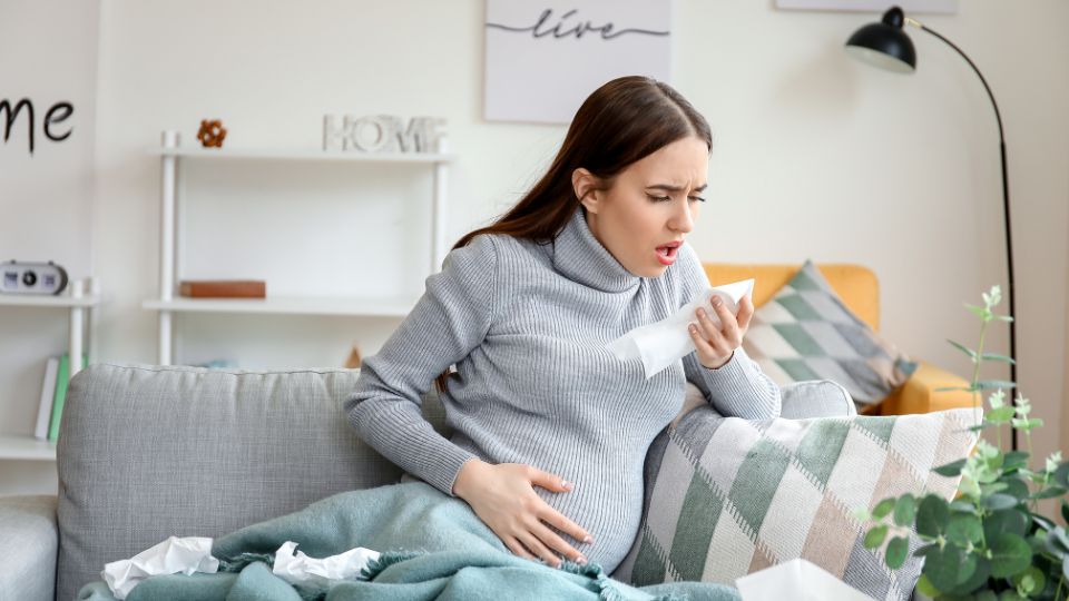 Pregnant woman sitting on a couch, holding her throat with a cup of tea nearby.