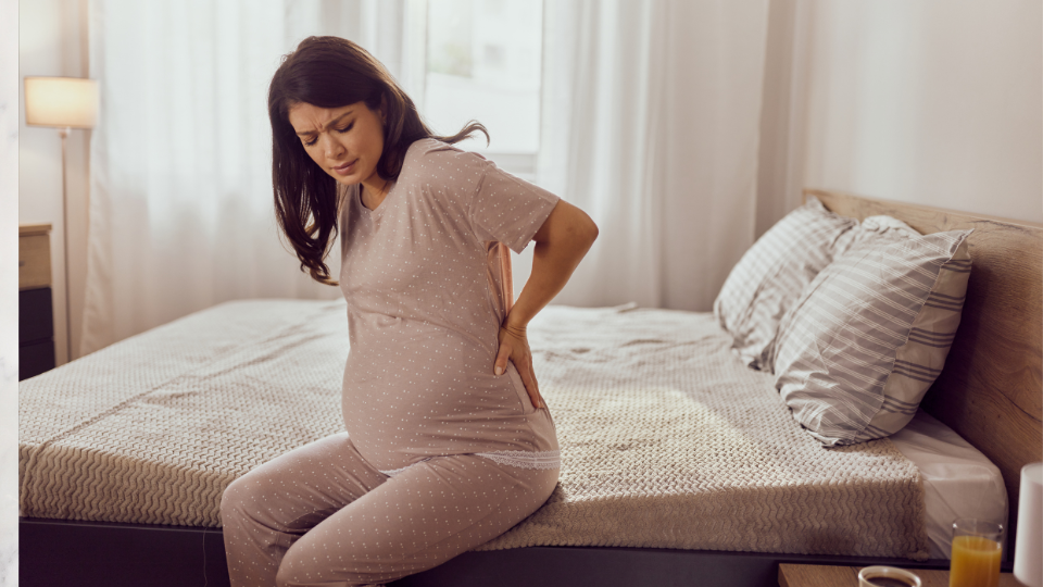 A pregnant woman holding her lower back and sitting on a comfortable chair, illustrating discomfort in the buttocks.