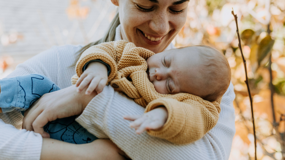 A mom gently holding a newborn in a cozy blanket while standing outside on a sunny day.